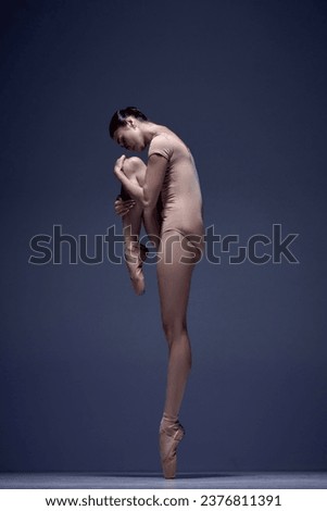 Elegant, attractive woman, professional ballerina in beige bodysuit, standing in pointe against blue studio background. Concept of classical dance, art and grace, beauty, choreography, inspiration
