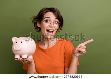 Photo of surprised young funny girl holding piggy bank directing finger novelty promoter invest money isolated on green color background