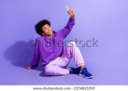 Full size photo of handsome person wear violet hoodie sitting making selfie on smartphone front camera isolated on purple color background