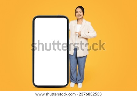 Happy chinese millennial lady in suit, point finger at big smartphone with empty screen, enjoy sale, ad and offer, isolated on orange studio background. Recommendation for work and business app
