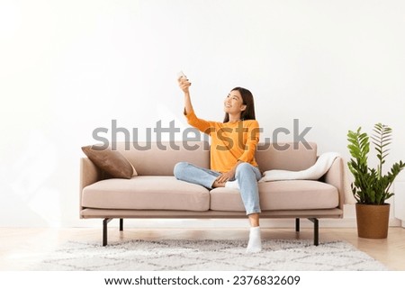 Happy pretty young asian woman wearing comfy casual outfit sitting on couch at home, holding remote control and looking at empty space over white empty wall, turning on AC