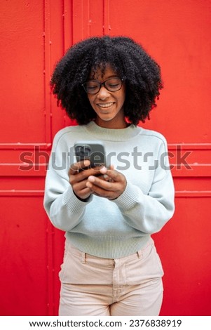 Portrait of a young african-american girl with afro hair holding her smartphone in her hands while laughing against a red background