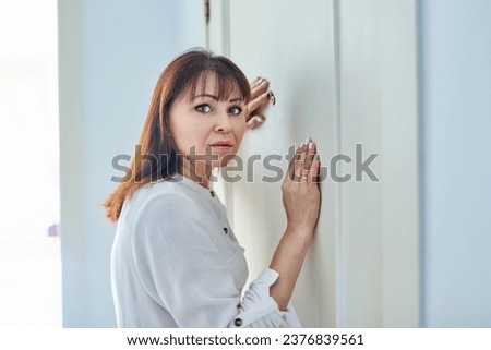 Serious middle-aged woman looking through peephole on front door Royalty-Free Stock Photo #2376839561