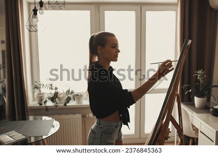A creative female artist stands and works on a painting. A happy young lady is painting on canvas in her apartment. The concept of a young mother's leisure