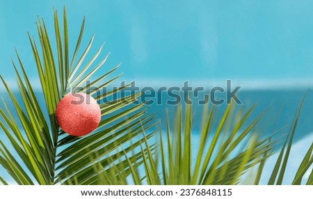 Christmas ball on palm tree branch on background of blue water pool at summer resort. Happy New Year and Merry Christmas on beach concept, celebration holidays in warm countries. Banner Copy space