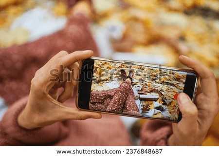 Close up hands holding smartphone and taking picture of her legs during picnic surrounded of orange leaves
