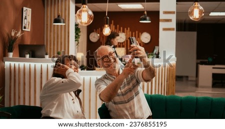 Elderly people taking photos in lobby, recording all activities on retirement vacation abroad. Old husband and wife take pictures on smartphone, making memories for family members.