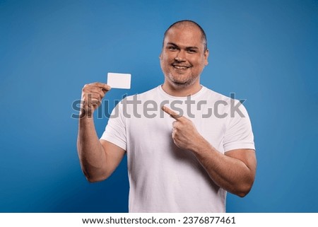 Portrait of a Brazilian man wearing a basic white shirt, facing forward, holding a paper card in his right hand, at chin height, with the index finger of his left hand pointing towards him, smiling