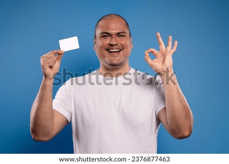 Portrait of a Brazilian man wearing a basic white shirt, facing forward, holding a paper card in his right hand, and making the OK sign with his left hand, smiling and looking at the camera - Belém