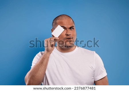 Portrait of a Brazilian man wearing a basic white shirt, from the front, holding a paper card in his right hand, covering one eye and looking at the camera - Belém - Pará - Brazil