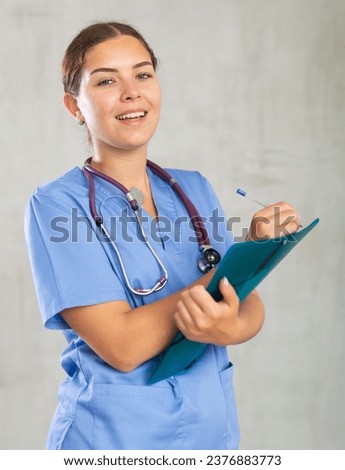 Portrait of young female medic in medical uniform making notes in folder in studio..