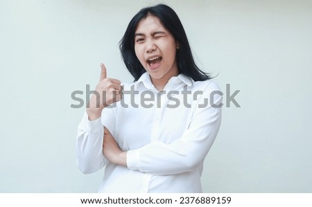 portrait of carefree satisfied asian young business woman manager giving thumbs up wearing white formal suit shirt, flirting blink eye to camera, show approve signs, standing over isolated background