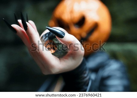 An adult model wearing a pumpkin head mask poses with a lens ball, which portrays her upside down; her hands are manicured with long, black press-on nails