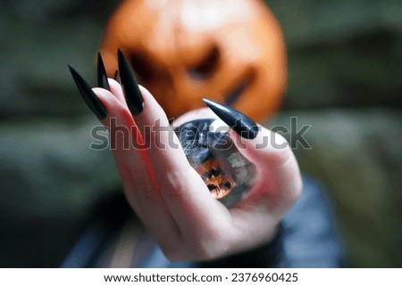 An adult model wearing a pumpkin head mask poses with a lens ball, which portrays her upside down; her hands are manicured with long, black press-on nails