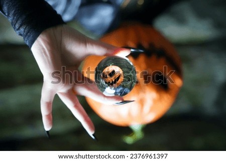 An upside-down view of an adult model wearing a pumpkin head mask poses with a lens ball, which portrays her right side up; her hands are manicured with long, black press-on nails