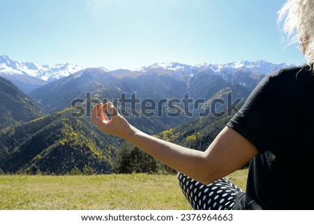 one, alone woman enjoys a sunny day outside in meadow while practising yoga. female girl doing meditation. Lotus pose. Rear back view of a lady meditating in nature. snow capped mountain on background