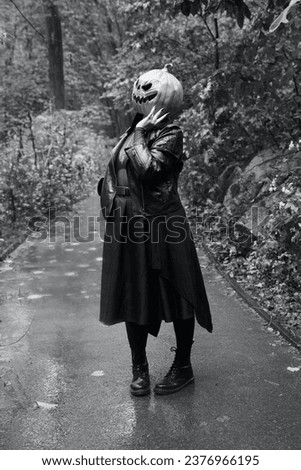 A black and white image of an adult model in a pumpkin head mask strolls down a walking path in Central Park in the rain