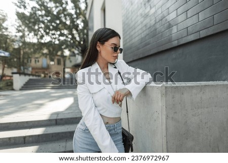 Female urban portrait of a beautiful fashion woman with vintage sunglasses in trendy stylish casual look outfit with a white blazer and blue jeans with a bag in the city