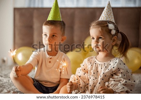Siblings are sitting surrounded by balloons with sprinklers in their hands and celebrating birthday.