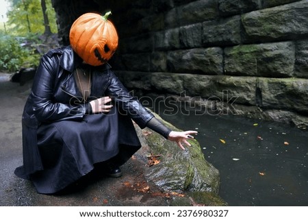 An adult model wearing a pumpkin mask squats next to The Loch stream and reachings out to the water with a manicured hand with long, black, press-on nails