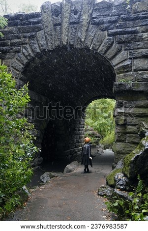 A wide shot of a model wearing a pumpkin head mask taking a stroll in the rain near Glen Span Arch in Central Park                               