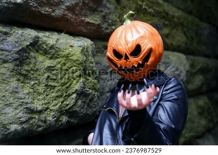 An adult model wearing a pumpkin head mask makes claw-like gestures to the camera with long, black, press-on nail
