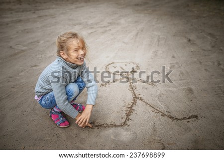 Little cheerful beautiful girl in sweater in sand draws funny man.