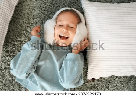 Cute little girl in warm earmuffs lying on carpet at home, top view Royalty-Free Stock Photo #2376996173