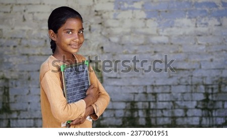 Portrait of happy cute little indian girl in school uniform holding blank slate, Adorable elementary kid showing black board. child education concept. rural india. Royalty-Free Stock Photo #2377001951