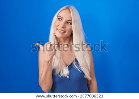 Caucasian woman standing over blue background smiling with happy face looking and pointing to the side with thumb up. 
