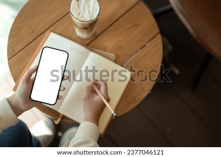 Top view image of a woman using her smartphone while keeping her diary or writing something in her book in a coffee shop. A white-screen smartphone mockup for display your graphic ads