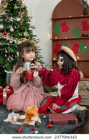Two pretty and joyful young Asian girls in cute Christmas dresses are enjoying Christmas hot chocolate and cookies and celebrating Christmas at home together. Friends, sibling, sister, special event