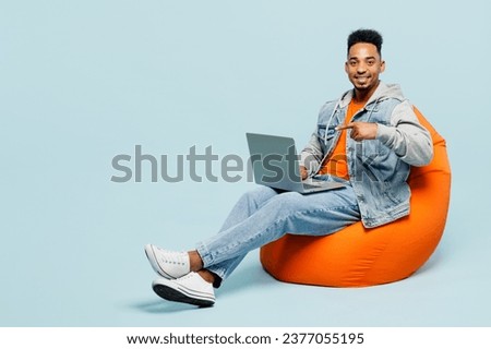 Full body young IT man of African American ethnicity wear denim jacket orange t-shirt sit in bag chair hold use work point finger on laptop pc computer isolated on plain pastel light blue background
