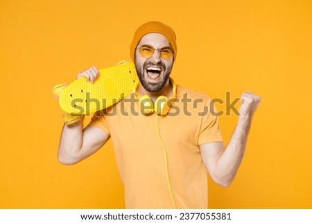 Happy screaming young bearded man 20s in basic casual t-shirt headphones eyeglasses hat hold skateboard doing winner gesture looking camera isolated on bright yellow colour background studio portrait