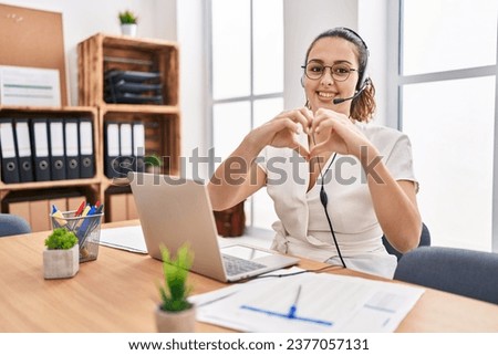 Young hispanic woman wearing call center agent headset at the office smiling in love showing heart symbol and shape with hands. romantic concept. 
