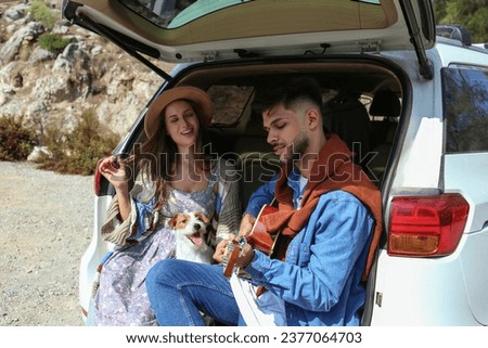 Happy hipster couple sitting in the trunk of the car and singing a song together with their rough coated jack russell terrier. Close up, copy space, background.