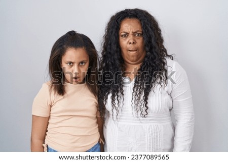 Mother and young daughter standing over white background in shock face, looking skeptical and sarcastic, surprised with open mouth 