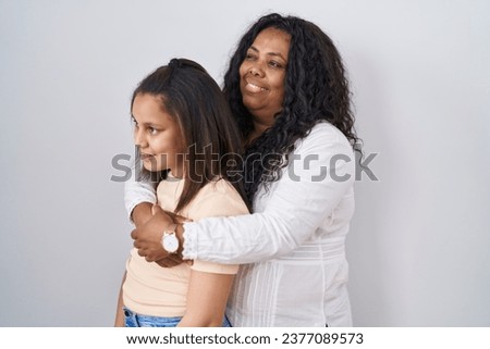 Mother and young daughter standing over white background looking to side, relax profile pose with natural face and confident smile. 