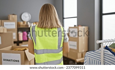 Attractive young blonde volunteer in reflective vest standing with her back to charity center, working at indoor warehouse, promoting community service and altruism. Royalty-Free Stock Photo #2377099311
