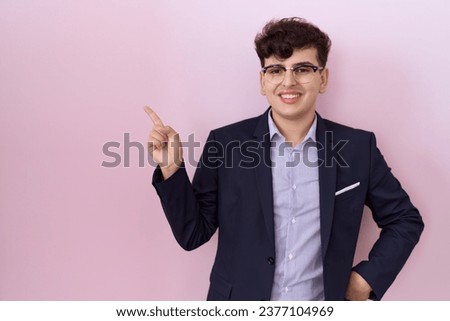 Young non binary man with beard wearing suit and tie with a big smile on face, pointing with hand and finger to the side looking at the camera. 