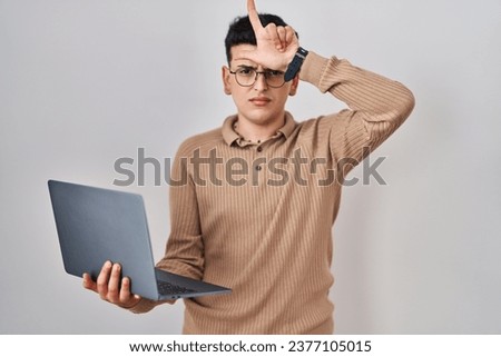 Non binary person using computer laptop making fun of people with fingers on forehead doing loser gesture mocking and insulting. 