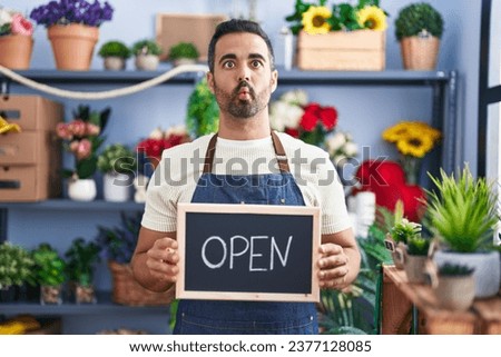 Hispanic man with beard working at florist holding open sign making fish face with mouth and squinting eyes, crazy and comical. 