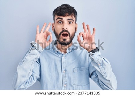 Young hispanic man with beard standing over blue background looking surprised and shocked doing ok approval symbol with fingers. crazy expression 