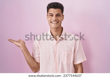 Young hispanic man standing over pink background smiling cheerful presenting and pointing with palm of hand looking at the camera. 
