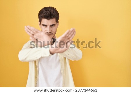 Young hispanic man standing over yellow background rejection expression crossing arms and palms doing negative sign, angry face 