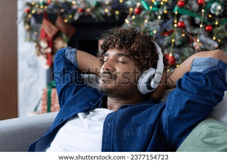 Indian young man wearing headphones, eyes closed and hands behind head sitting on sofa at home and resting, dreamy and relaxed listening to music on New Year and Christmas holidays. Close-up photo.