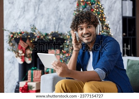 Portrait of a young hispanic man sitting at home on the sofa near the Christmas tree during the winter holidays. Wearing headphones, he listens to music from a tablet, rests, smiles at the camera. Royalty-Free Stock Photo #2377154727