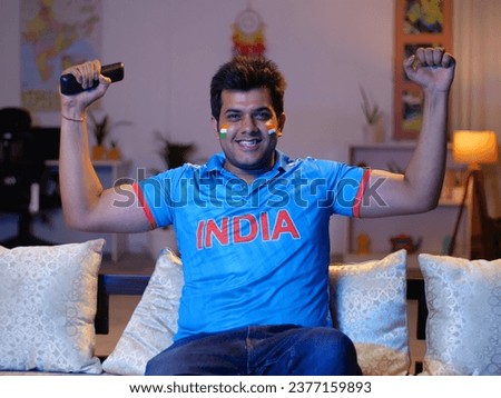 Excited young Indian man wearing cricket jersey - watching cricket match on TV, excitement. Indian man enthusiastically making supporting and winning gestures while watching a live cricket match on...