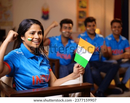 Bokeh shot of cricket fans watching a live match at home - a female fan holding an Indian flag in her hands, cheering up for team India. A group of friends huddled together- wearing a jersey