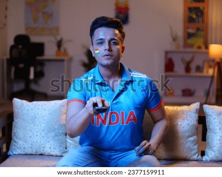 A young Indian man wearing a cricket jersey - watching a cricket match on TV, young cricket fan. An Indian man watching a live cricket match at home - wearing a jersey to support their team India